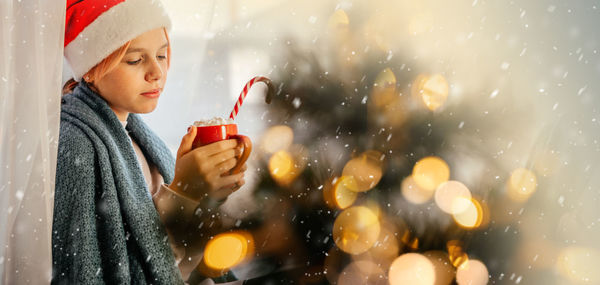 Young teen girl in santa hat having a cup of hot chocolate with marshmallow with christmas tree on
