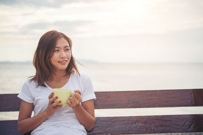 Smiling woman holding coffee cup while sitting on wooden swing at beach against sky during sunset