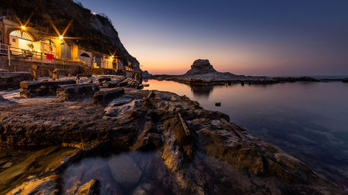 Scenic view of sea by illuminated buildings against sky during sunset