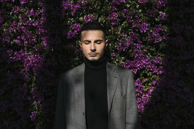 Fashionable man standing with eyes closed against flowering plant wall