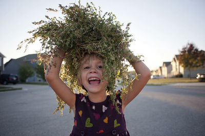 Portrait of happy girl carrying plants on head while walking at street against sky