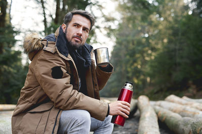 Young man drinking drink sitting in forest