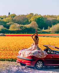 Woman sitting on car by flowering plants against sky