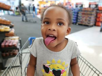 Portrait of cute girl sticking out tongue while sitting in shopping cart at supermarket