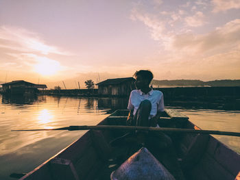 Rear view of man sitting on lake against sky during sunset