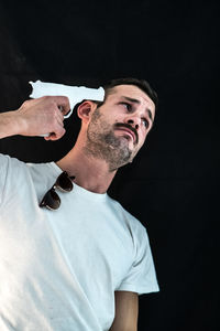 Low angle view of depressed man holding gun on head against black background