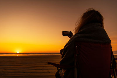Rear view of silhouette woman photographing at beach against sky during sunset