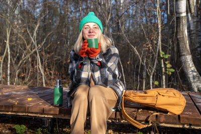 Relaxed woman hiker heating drinking hot tea sitting on wooden path in autumn park in scandinavia.