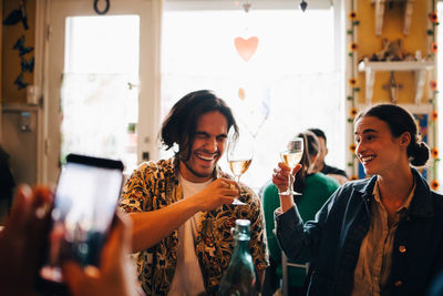 Man photographing cheerful young friends raising celebratory toast at restaurant during brunch