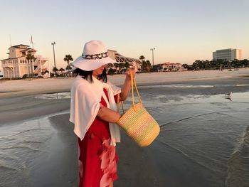 Side view of woman holding shoulder bag while standing at beach sky during sunset