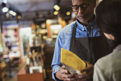 Owner reading label on food packet while standing with customer at store