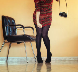 Low section of young woman adjusting dress while holding camera against wall