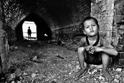 Portrait of boy sitting against brick wall with brother standing in background