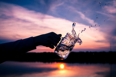 Cropped hand pouring water from jar against sky during sunset