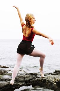 Full length of ballet dancer practicing on rocks at beach against clear sky