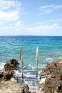Steel ladder on rocky coastline in sea against sky
