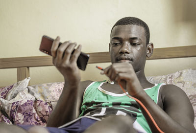 Focused young black male with short hair lying on bed and plugging usb cable into mobile phone