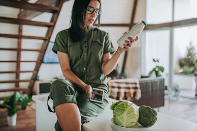 Woman with milk bottle using mobile phone in kitchen at home