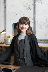 Portrait of confident smiling young female owner standing against window in shop