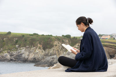 Side view of female in casual clothes reading book near sea