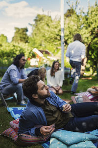 Young man eating while relaxing in backyard with friends in background