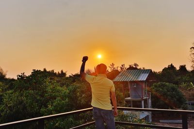 Rear view of man standing by railing against sky during sunset