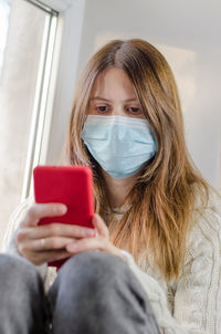 Woman in a medical mask sitting on the windowsill looking into a smartphone