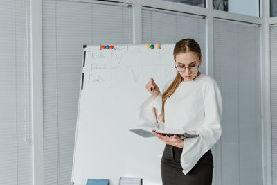 Businesswoman reading diary while standing by whiteboard in office