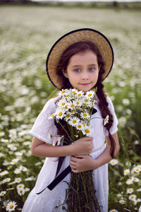 Portrait girl child in a white dress stands on a camomile field in a hat. bouquet of flowers