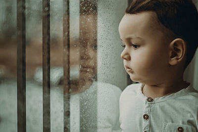 Portrait of cute boy looking through wet window