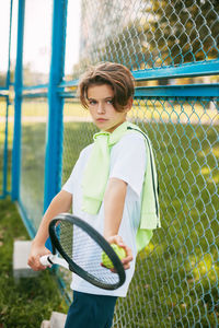 Portrait of boy with tennis racket standing by fence