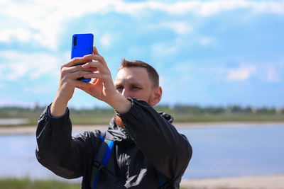 Bearded man in black raincoat on blue sea landscape background with smartphone in hands takes selfie