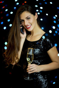Portrait of beautiful young woman holding champagne flute while talking on phone against illuminated lights