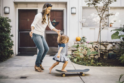 Daughter balancing over skateboard with help of mother on footpath