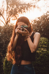 Portrait of woman holding camera standing against trees