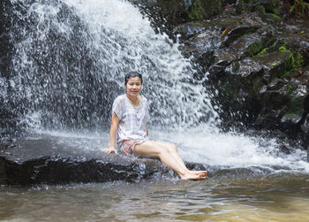 Portrait of smiling woman sitting on rock against waterfall