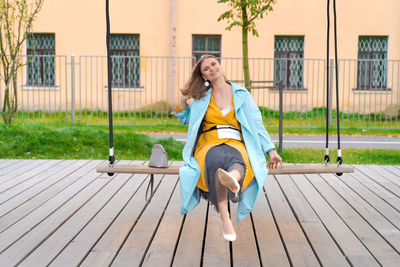 Happy woman swinging on swing in city park, wearing yellow dress and blue coat