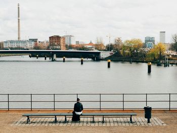 Rear view of woman sitting on bench by river in city