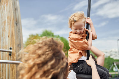 Happy adorable little girl riding rope swing while having fun on playground under supervision of mother