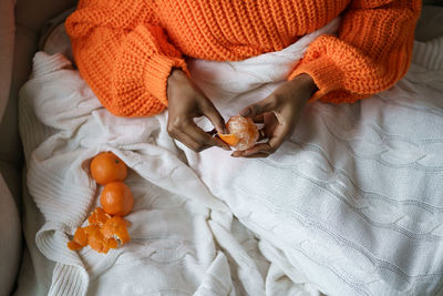 Afro woman hands peeling ripe sweet tangerine, wear orange sweater, lying in bed under the blanket