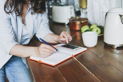 Adult smiling brunette woman in casual doing notes in daily book with cup of tea on kitchen at home