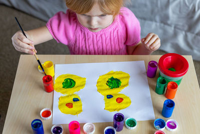 Little smiling girl painting on paper with colorful paints sitting at the table at home