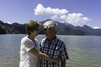 Happy senior couple embracing by lake against sky