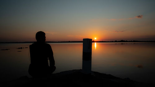 Silhouette man sitting on pier by sea against sky during sunset