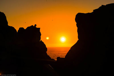 Silhouette rocks by sea against orange sky