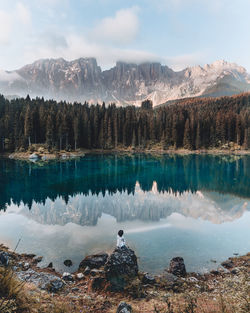 Rear view of woman sitting on rock by lake against sky during winter