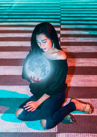 Digital composite image of woman holding illuminated moon while sitting on road