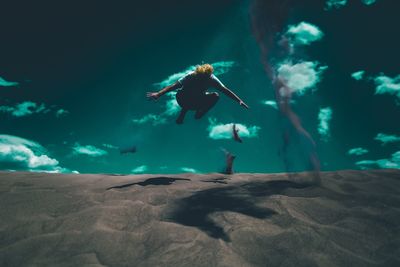 Low angle view of playful man jumping while throwing sand at beach