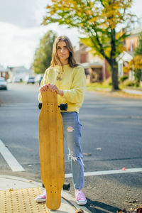 Girl standing on corner of street holding longboard in fall colors