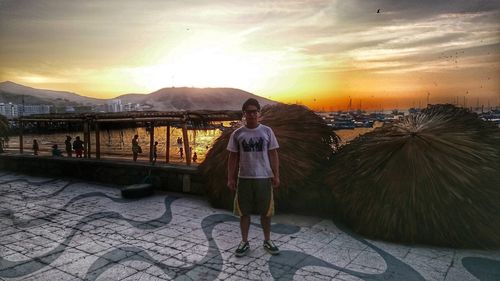 Portrait of man standing on promenade at beach against orange sky during sunset